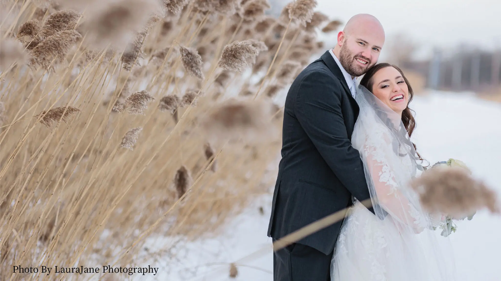 Wedding couple posing outside in the snow next to blooming cattail plants.