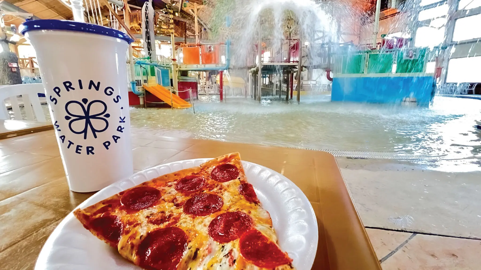 Close up of a Springs Water Park souvenir cup and slice of pizza with water park play structure in the background.