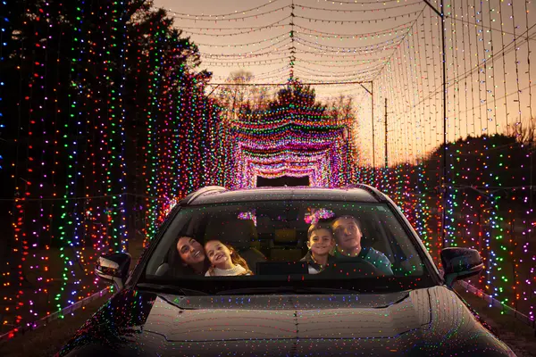 A family of four smiling and looking out the windshield of a car at Christmas lights.