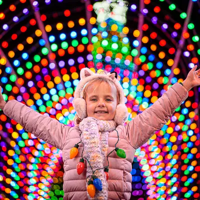 A girl with her arms extended smiling in a Christmas light tunnel.