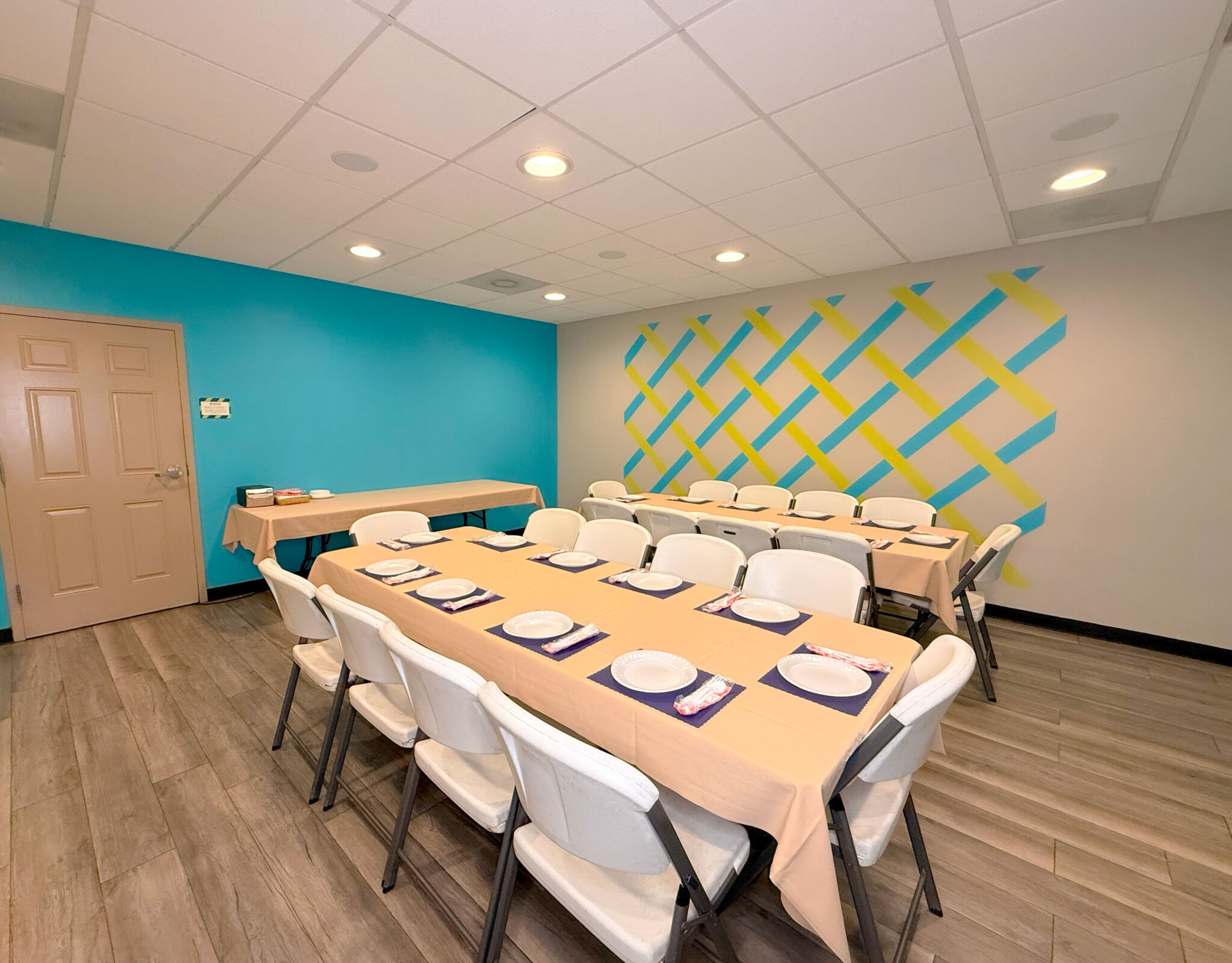 The teal lower party room rectangular table, cream tablecloths, white chairs, and a teal and lime wall mural.
